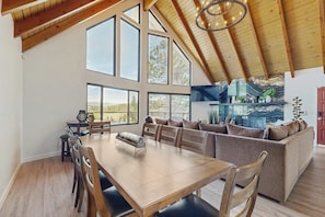 A spacious dining area with a wooden table and chairs, a large window, and a modern kitchen with a stone backsplash. - Modern chalet w/expansive deck, gas grill, & game room - close to lake & town (Lake Arrowhead)