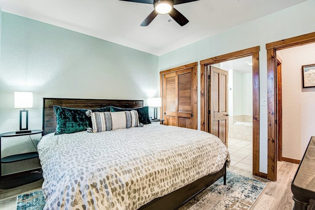 A bedroom with a bed, a ceiling fan, a nightstand, and a door leading to another room.