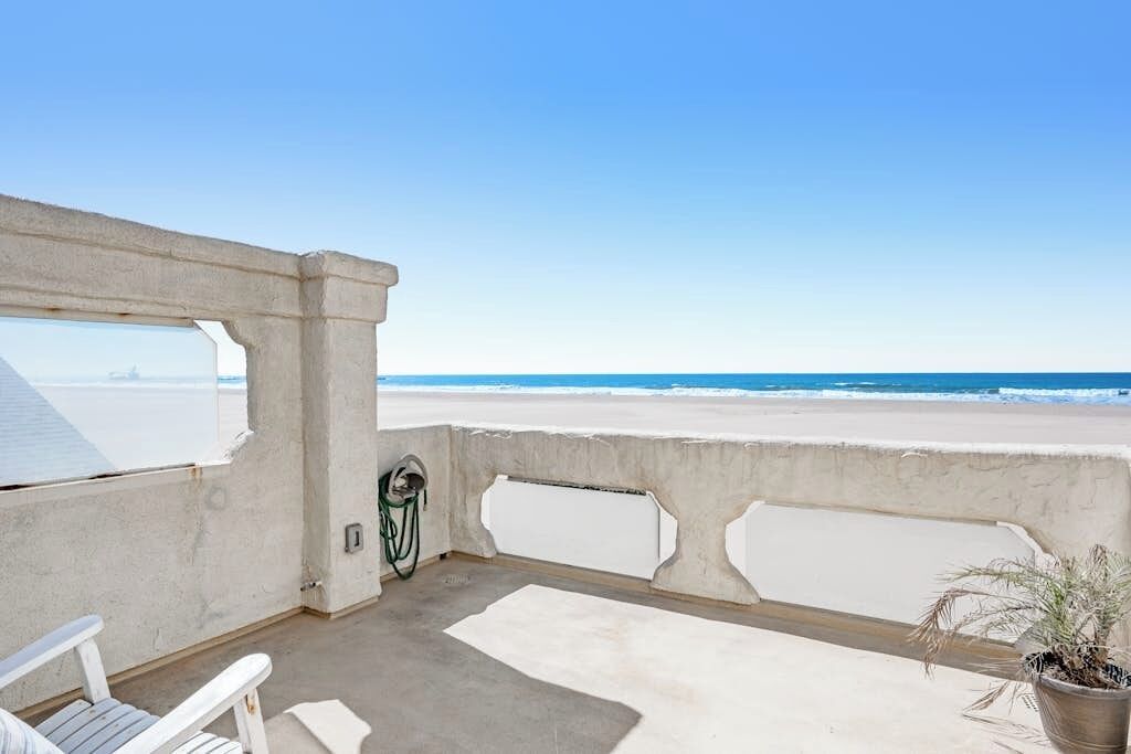 A balcony with a view of the beach, a chair, a potted plant, and a power outlet.