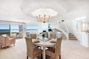 A spacious living area with a large round table, wicker chairs, a chandelier, and a view of the ocean. - Oceanfront Home with Giant Picture Windows, Gas Fireplace, and Free WiFi (Oxnard)