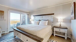 A bedroom with a bed, a nightstand, a lamp, and a view through a glass door. - Stunning ocean-view home with W/D, sauna, tennis court, & patio - beach access (Moss Landing)