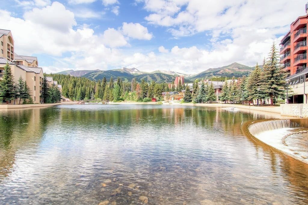 Un lago con edificios a la izquierda y a la derecha, rodeado de árboles y montañas al fondo.
