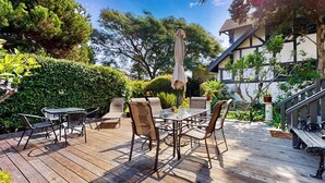 A wooden deck with outdoor furniture, including a table and chairs, surrounded by greenery and a building with a balcony. - Classic beach suite 1 block to ocean - fireplace, lovely patio, near downtown (Santa Barbara)