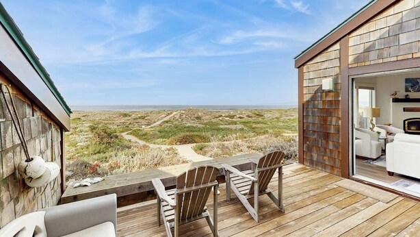 A wooden deck with outdoor furniture, a view of the beach, and a building with a glass door.