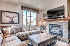 Ein Wohnzimmer mit Ecksofa, Kamin, Fernseher und großem Fenster mit Blick auf die Stadt.