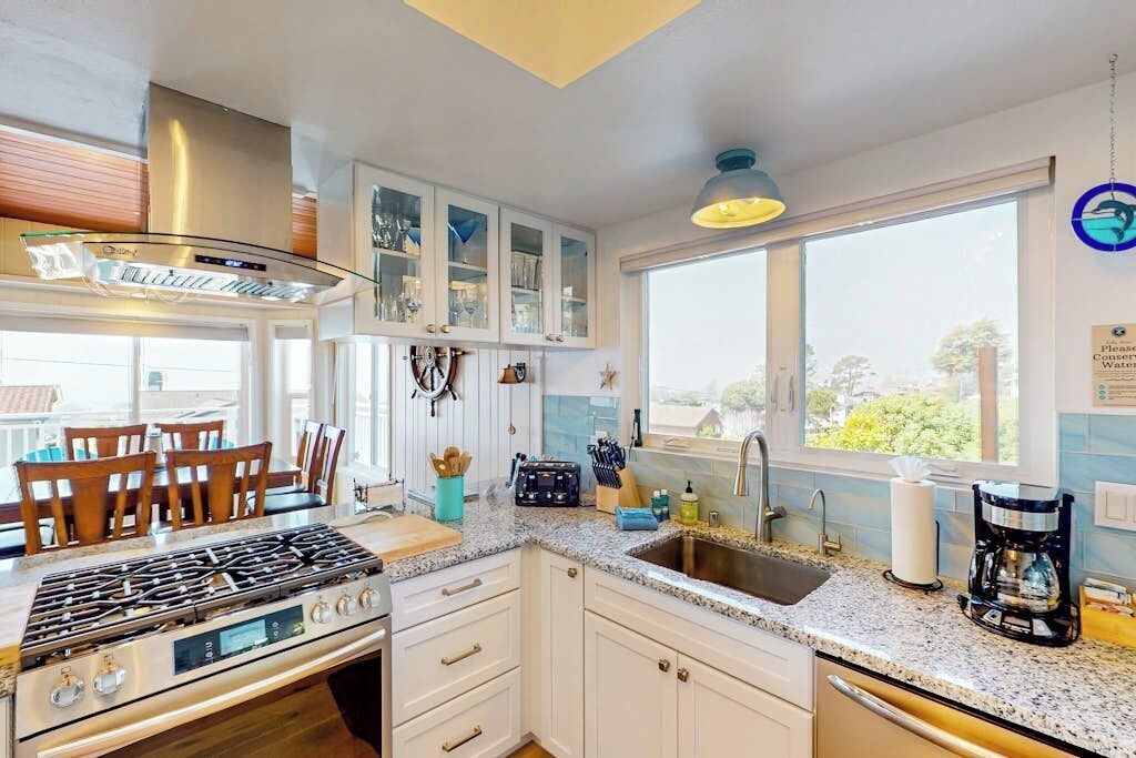 A modern kitchen with a gas stove, white cabinets, a large window, and a dining area with wooden chairs.