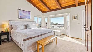A bedroom with a large bed, a bench, a nightstand, a window, and a door. - Panoramic Ocean and Sunset Views with EV Charging available (Cambria)
