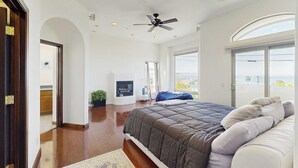 A bedroom with a large bed, a ceiling fan, a fireplace, and a view of the outdoors.