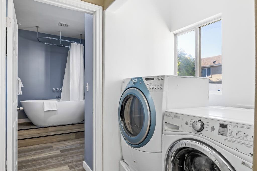 A laundry room with a front-loading washing machine, a bathtub, and a window providing natural light.