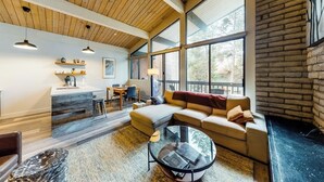 Una sala moderna con techo de madera, ventanas amplias, un sofá, una mesa de centro y una cocina con barra y taburetes.