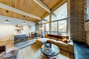 Ein modernes Wohnzimmer mit Holzboden, großen Fenstern, einer Couch, einem Couchtisch und einer Küchenzeile mit einer Arbeitsplatte und Hockern.