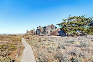 A modern house with a pathway leading to it, surrounded by dry vegetation and a clear sky. - Beachfront home with heated pool, hot tub, sports courts, amazing views, & WFH (Moss Landing)