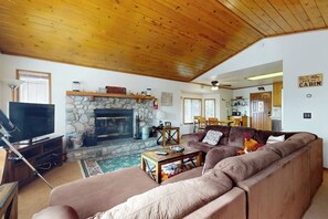 A cozy living room with a stone fireplace, wooden ceiling, and a large brown sofa. - Cheery cabin with gas grill, patio, deck, outdoor space, & near lake (Lake Arrowhead)