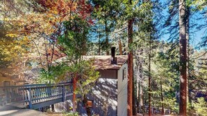 A rustic cabin with a wooden deck and a metal railing, surrounded by tall trees and a clear sky. - Mountainside Retreat w/ Fireplace, BBQ & Furnished Deck - Near Village (Lake Arrowhead)