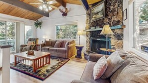 A spacious living room with a stone fireplace, wooden ceiling beams, and large windows. - Mountainside Retreat w/ Fireplace, BBQ & Furnished Deck - Near Village (Lake Arrowhead)