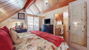 A bedroom with a sloped ceiling, a bed with a floral bedspread, a dresser, a television, and a door with a glass panel. - Cabin w/ Loft, hot tub, Fireplace, game room & Private lake trail access (Lake Arrowhead)