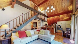 A spacious living area with a staircase, a sofa, a lamp, and a kitchen in the background. - Cabin w/ Loft, hot tub, Fireplace, game room & Private lake trail access (Lake Arrowhead)
