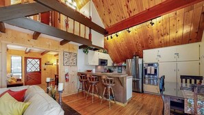 A rustic cabin interior with a wooden ceiling, a kitchen with bar stools, and a dining area. - Cabin w/ Loft, hot tub, Fireplace, game room & Private lake trail access (Lake Arrowhead)