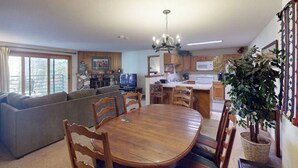 A dining area with a wooden table and chairs, a sofa, a television, and a kitchen in the background. - Conveniently Located Condo with Shared Pool, Hot Tub - Walk to Slopes (Keystone)