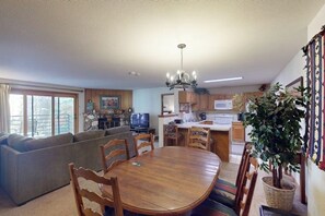 A dining area with a wooden table and chairs, a sofa, a television, and a kitchen in the background. - Conveniently Located Condo with Shared Pool, Hot Tub - Walk to Slopes (Keystone)