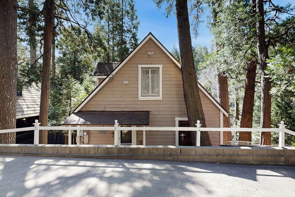 A single-story cabin with a gabled roof, surrounded by tall trees, and a white picket fence in front.