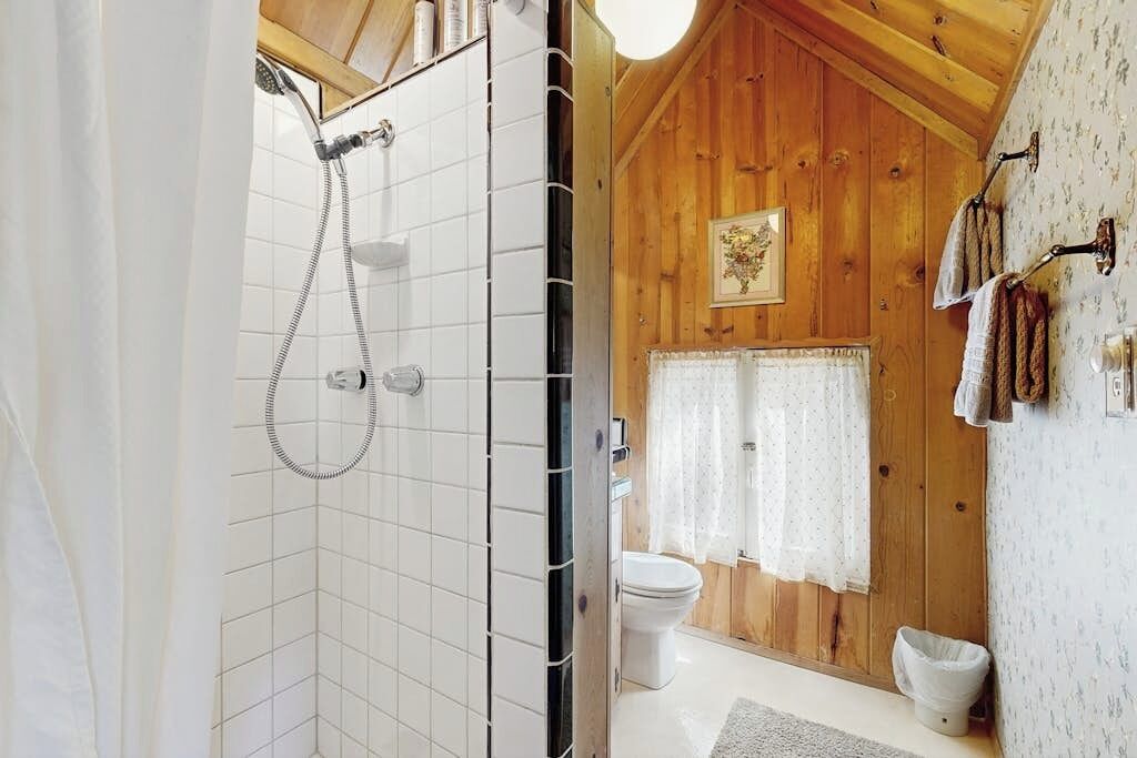 A wooden cabin bathroom with a shower, toilet, and a window with curtains.