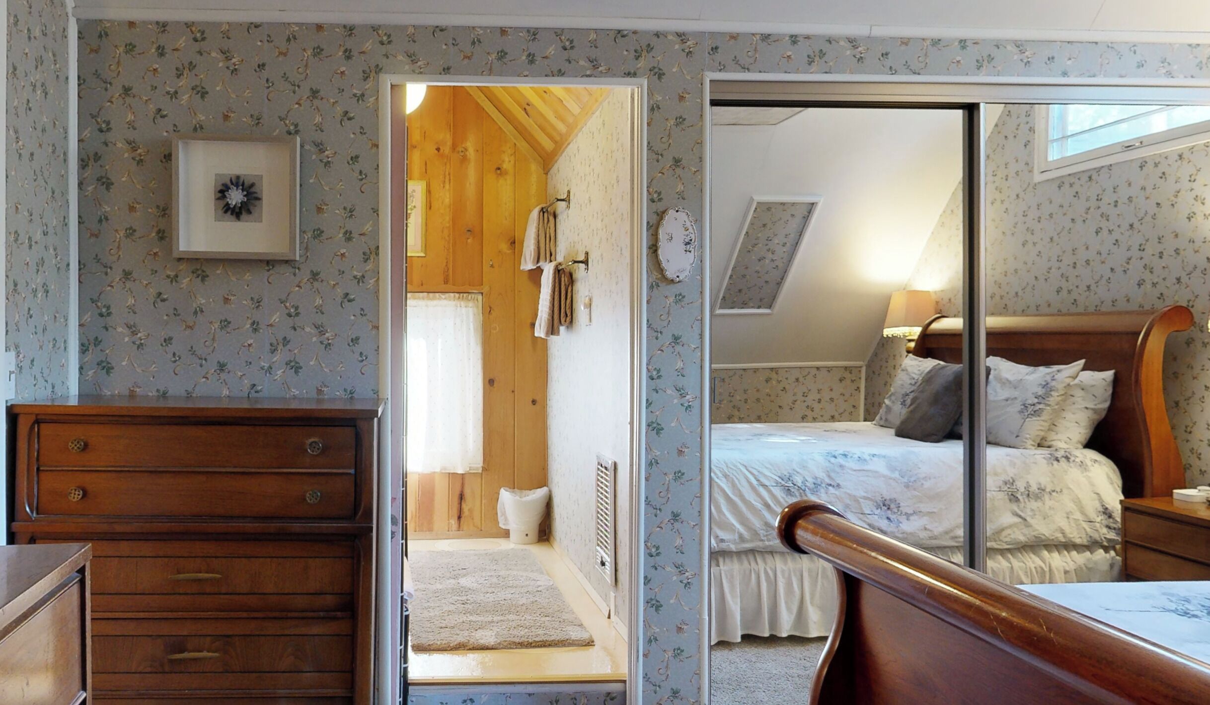 Une chambre à coucher comprenant un lit, une commode en bois, un miroir et une porte donnant sur une salle de bain.