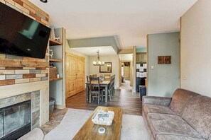 Ein Wohnzimmer mit Kamin, Fernseher, einer Couch, einem Essbereich mit Tisch und Stühlen sowie einer Küche im Hintergrund.
