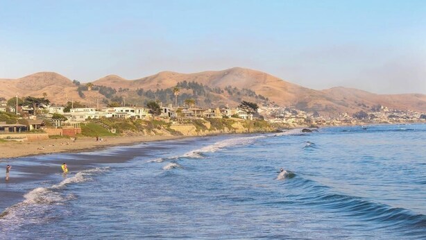 A coastal view with a beach, ocean waves, and a town in the background. - Classic getaway by the sea within walking distance to everything w/ocean views (Cayucos)