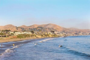 A coastal view with a beach, ocean waves, and a town in the background. - Classic getaway by the sea within walking distance to everything w/ocean views (Cayucos)