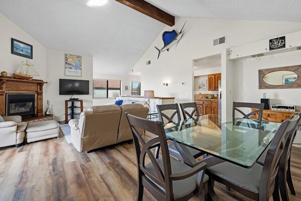 A spacious living area with a glass dining table, a fireplace, a television, and a kitchen in the background.