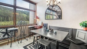 Ein Essbereich mit einem großen Tisch, Stühlen, einer Chandelierleuchtre und Blick auf einen Wald durch große Fenster.