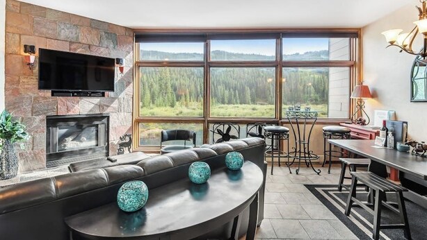 Ein modernes Wohnzimmer mit Kamin aus Stein, einem großen Fernseher und Blick auf einen Wald durch große Fenster.