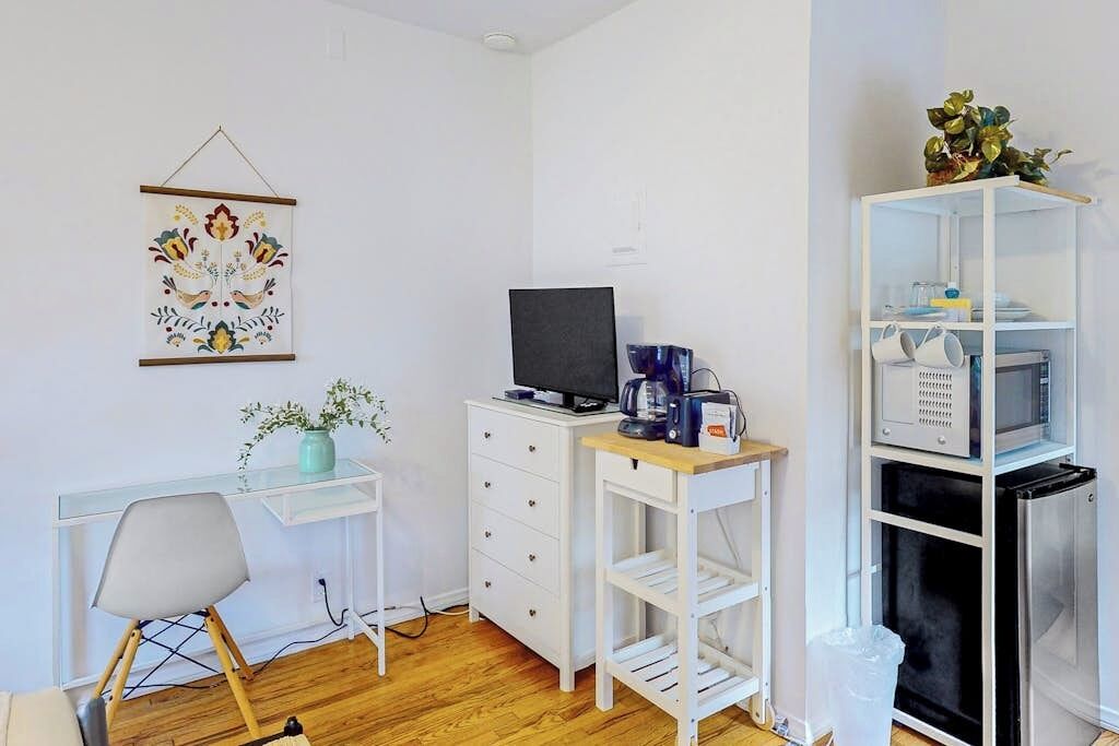 Une pièce comprenant un bureau, une chaise, une télévision, une machine à café et une étagère contenant des ustensiles de cuisine.
