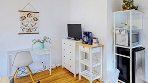 A room with a desk, chair, television, coffee machine, and a shelving unit with kitchen items.