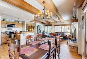 A spacious kitchen with wooden beams, a dining area with a large wooden table, and a cozy living area with a sofa and fireplace. - Cozy Condo with Stunning Mountain Views, Hot Tub, and Fireplace (Crested Butte)