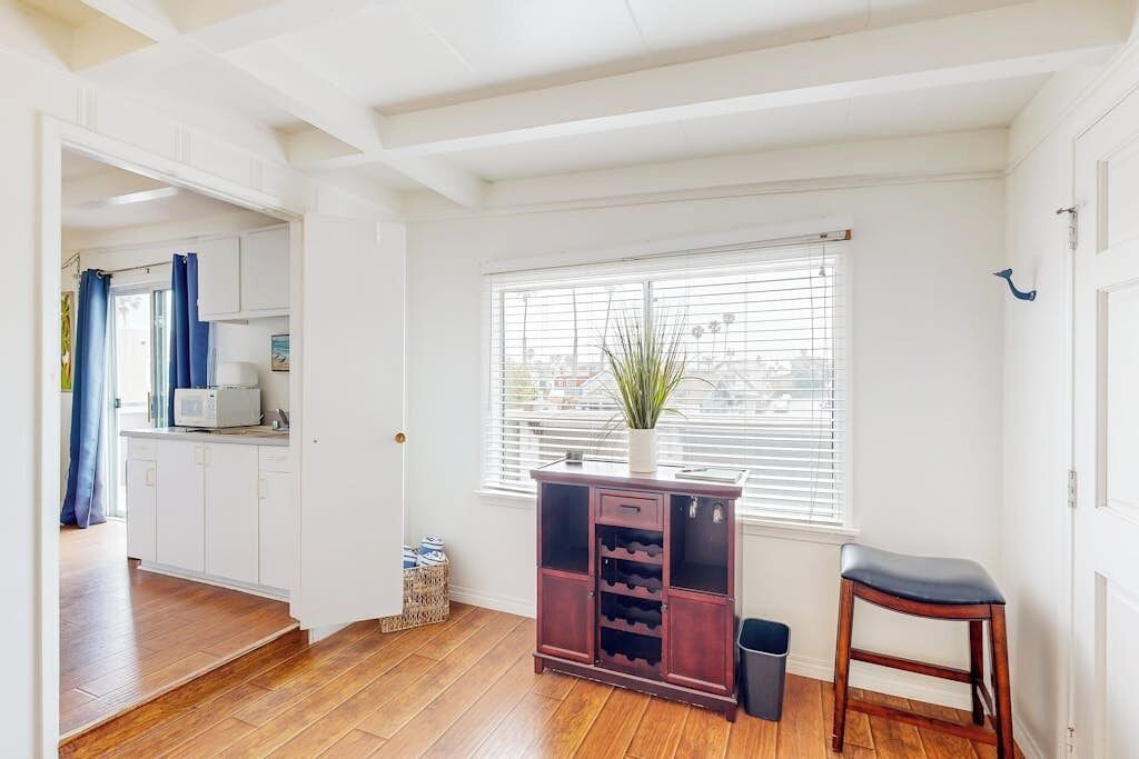 A room with a wooden floor, a window with blinds, a white door, and a small cabinet with wine bottles.