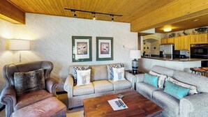 Una sala de estar con sillones de cuero, un sofá y una mesa de centro. Se observan cuadros enmarcados en la pared y una zona de cocina al fondo.
