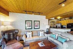 Ein Wohnzimmer mit Ledersesseln, einer Couch und einem Couchtisch. An den Wänden hängen gerahmte Bilder, und im Hintergrund ist eine Küchenzeile zu sehen.