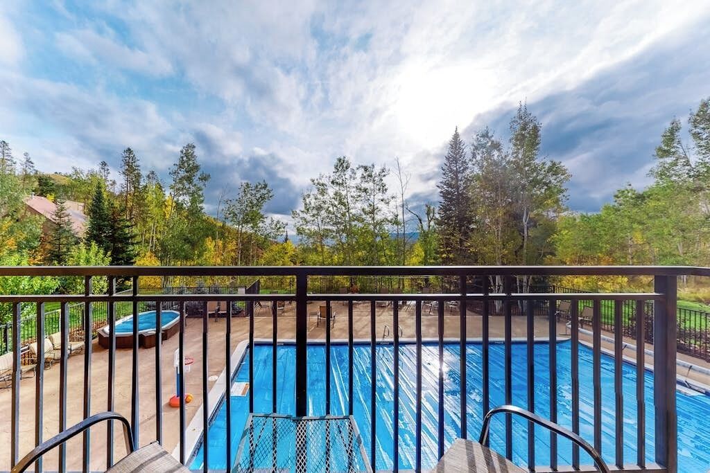A swimming pool with a metal railing, surrounded by trees and a cloudy sky.