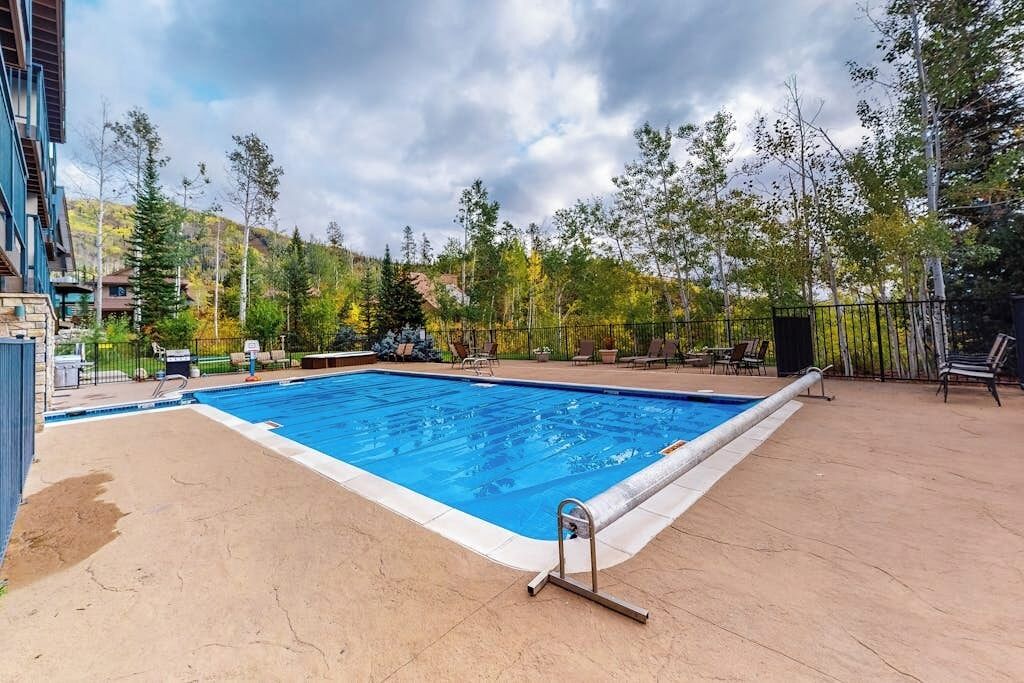 A rectangular swimming pool with a ladder, surrounded by a patio area with chairs and tables.