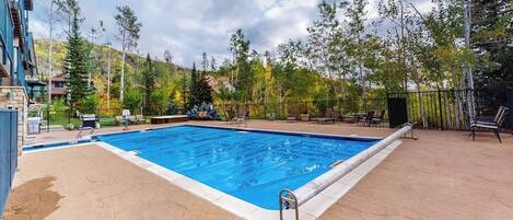 A rectangular swimming pool with a ladder, surrounded by a patio area with chairs and tables.
