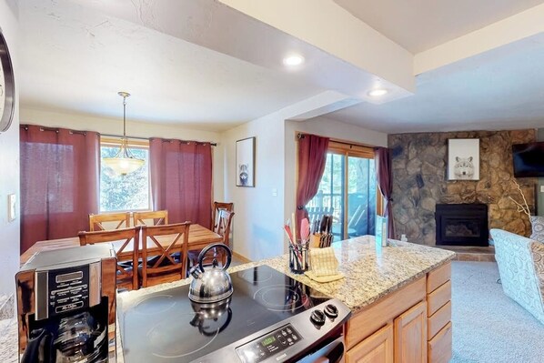 A kitchen with a stove, a kettle, a coffee maker, a dining table with chairs, and a stone fireplace. - Mountain view condo with hot tub, walk to Snowflake Lift & Main Street (Breckenridge)