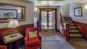 Un vestíbulo de hotel con un sillón rojo, una mesa de madera con una lámpara, una escalera y cuadros enmarcados en las paredes.