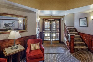 Eine Hotelhalle mit einem roten Sessel, einem Holztisch mit Lampe, einer Treppe und gerahmten Bildern an den Wänden.