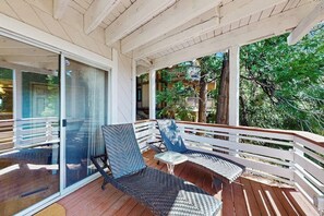 Ein überdachter Außenbereich mit Korbstühlen und einem kleinen Tisch, eine Glastür, die auf einen Balkon führt, und eine Holzterrasse.