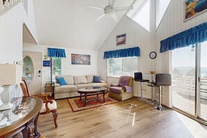 Ein großzügiges Wohnzimmer mit hoher Decke, einem Esstisch mit Stühlen, einer Sofagarnitur, einem Couchtisch und Blick durch große Fenster.