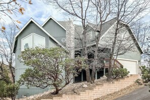A two-story house with a stone chimney, a covered porch, and a staircase leading to the garage. - Multi-level home with pool table, AC, fireplaces, balcony & gorgeous views (Lake Arrowhead)