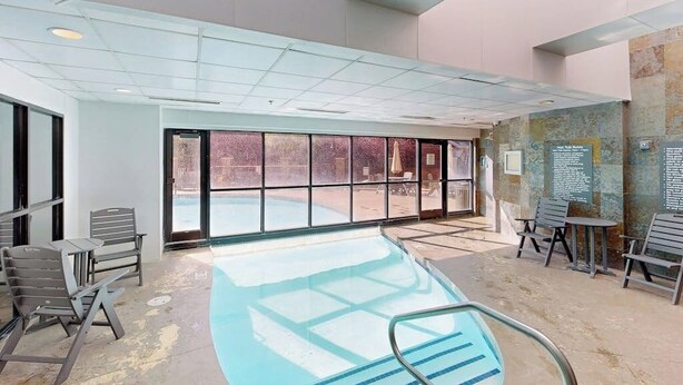 Una piscina interior con paredes de vidrio transparente, suelo de baldosas y varias sillas y mesas de descanso.