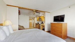 Un dormitorio con cama, televisor, cómoda de madera y una cocina visible a través de una puerta abierta.